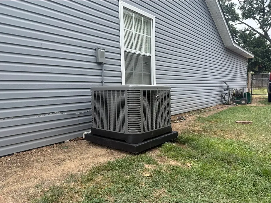New heat pump condenser on a black pad beside grey siding with a lawn in the foreground